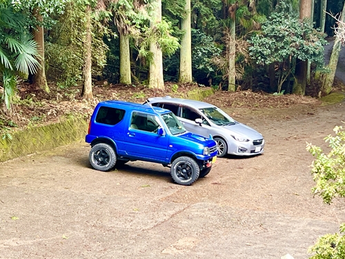 豊かな緑に囲まれた駐車場に並ぶ青いジムニーJB23とシルバーのスバル・インプレッサ。