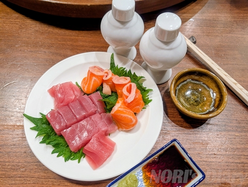 白い皿に盛り付けられた新鮮なマグロとサーモンの刺身。大葉が敷かれ、手前には醤油とわさび、奥には徳利とお猪口が並ぶ晩酌の風景。