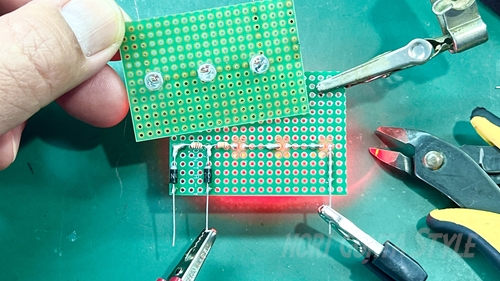 緑色のユニバーサル基板上で、3つのLEDをハンダ付けしている様子。基板はクリップで固定され、ニッパーなどの工具が置かれている。