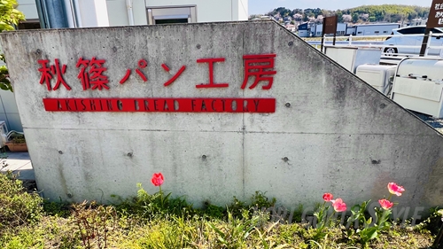 「秋篠パン工房」と赤い切り文字で書かれたコンクリートの門柱。足元には赤いストロベリーキャンドルが咲いている。