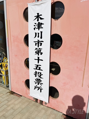 木津川市第十五投票所と書かれた白い垂れ幕が、丸い窓のあるピンク色の建物の入り口に掲げられている風景
